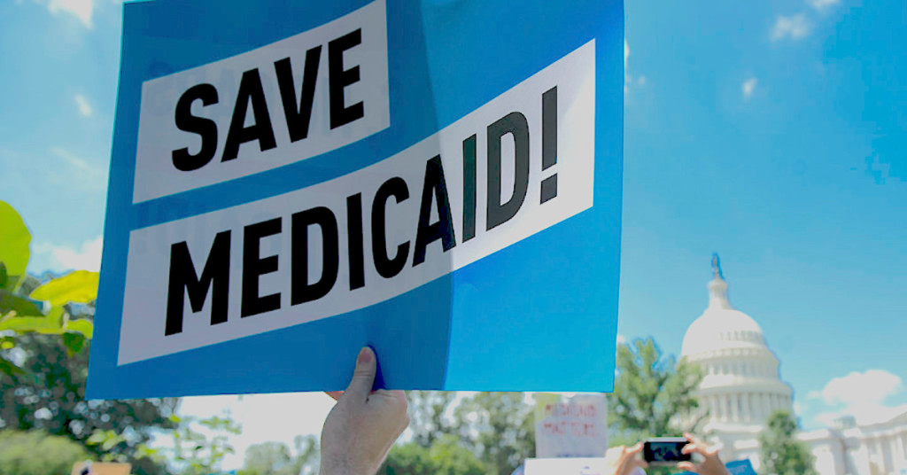 Hand holding up a blue sign saying "Save Medicaid!" with Capitol building in background